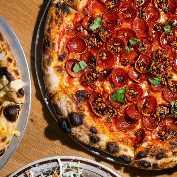 Close-up of a pepperoni pizza with chili crisp and basil, alongside other dishes on a wooden table.