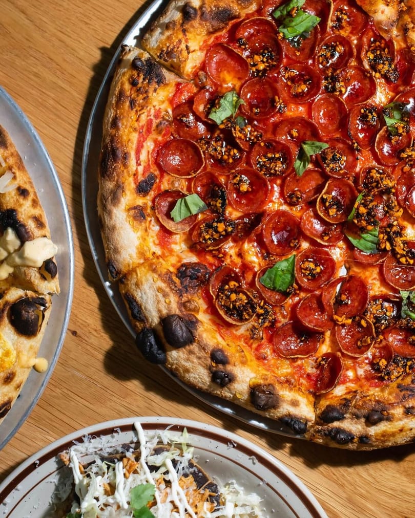 Close-up of a pepperoni pizza with chili crisp and basil, alongside other dishes on a wooden table.
