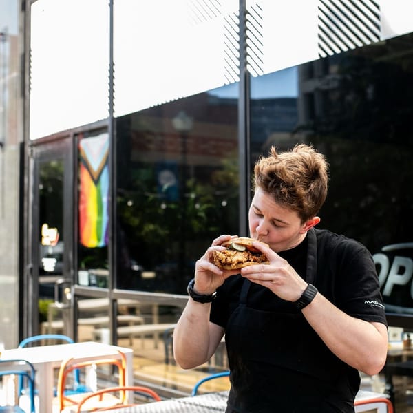 A person in a black apron takes a bite of a fried chicken sandwich outside a restaurant.