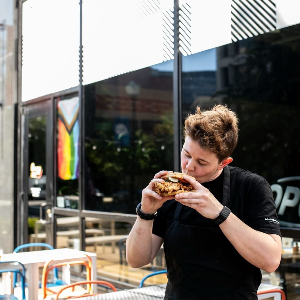 A person in a black apron takes a bite of a fried chicken sandwich outside a restaurant.