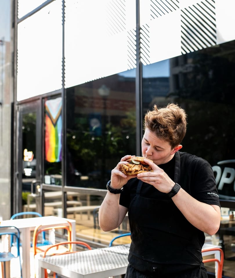 A person in a black apron takes a bite of a fried chicken sandwich outside a restaurant.
