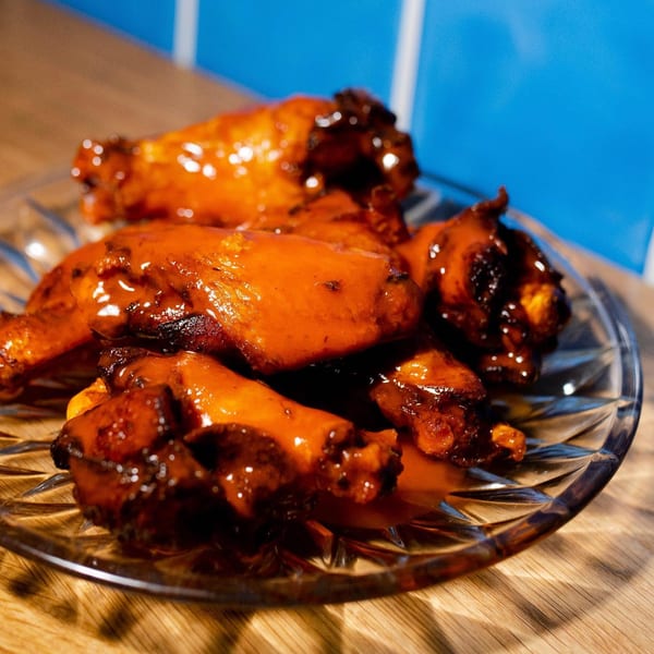 A glass plate of glistening, saucy chicken wings on a wooden surface with a blue tiled background.