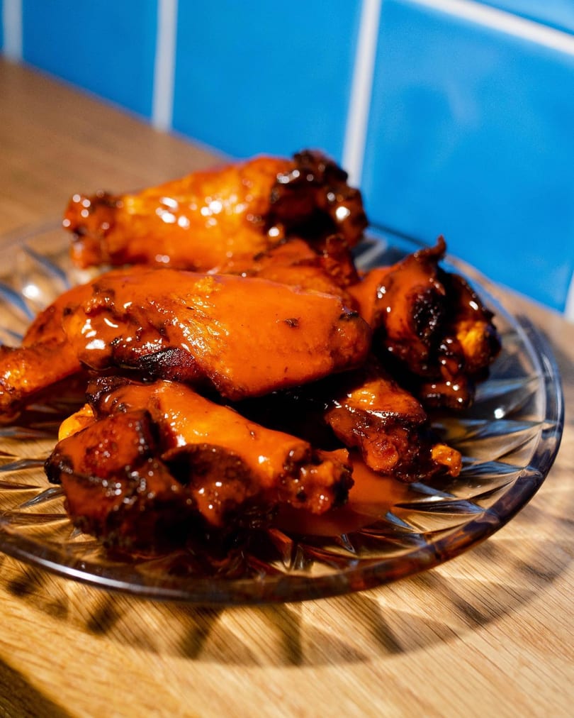 A glass plate of glistening, saucy chicken wings on a wooden surface with a blue tiled background.