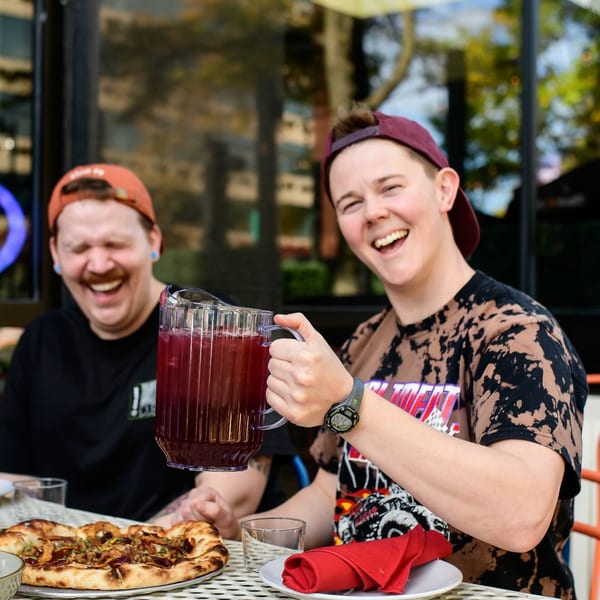 Two laughing people enjoying pizza and a pitcher of red drink on an outdoor patio.