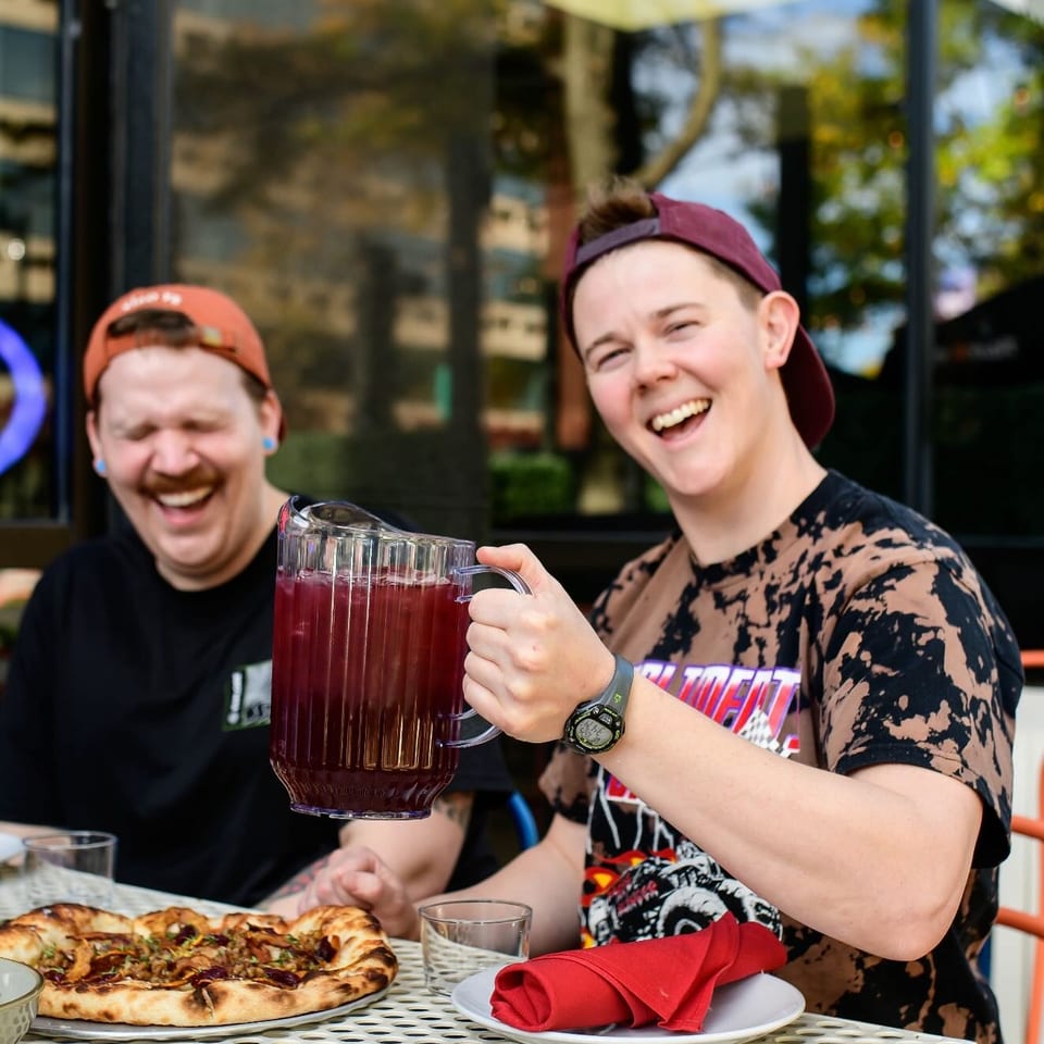 Two laughing people enjoying pizza and a pitcher of red drink on an outdoor patio.