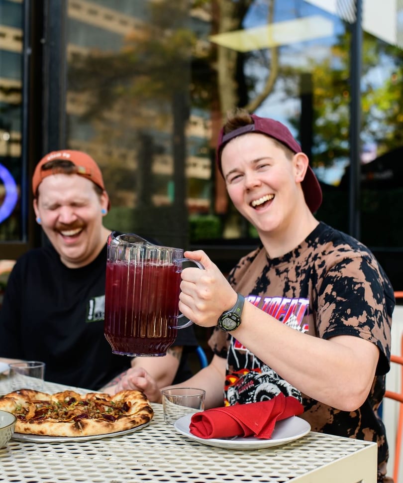Two laughing people enjoying pizza and a pitcher of red drink on an outdoor patio.
