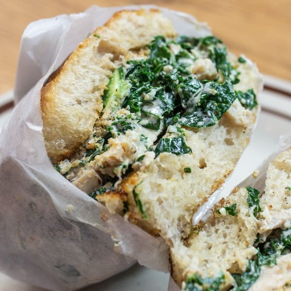 A large sandwich with creamy green filling, wrapped in white paper, cut in half on a plate.