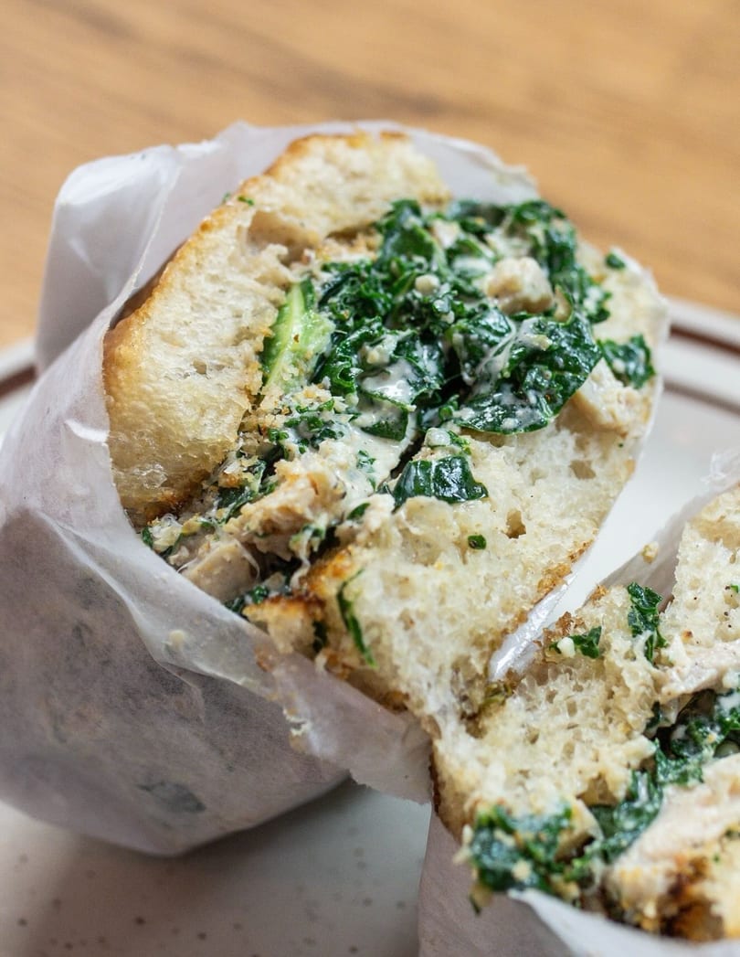 A large sandwich with creamy green filling, wrapped in white paper, cut in half on a plate.