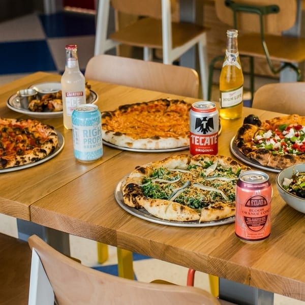 A wooden table laden with four pizzas, a side dish, and various canned and bottled drinks.