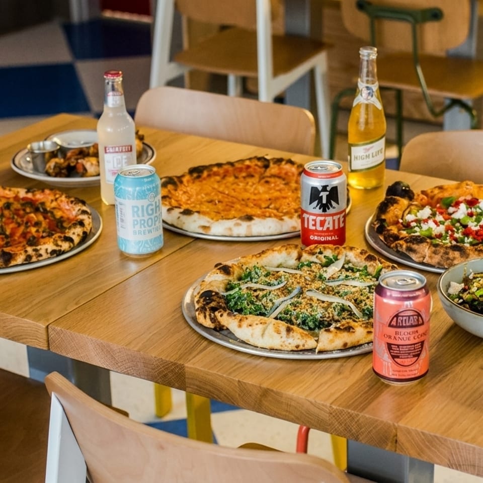 A wooden table laden with four pizzas, a side dish, and various canned and bottled drinks.