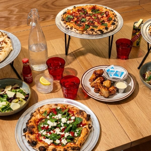 A wooden table laden with three pizzas, two salads, chicken wings, and drinks.