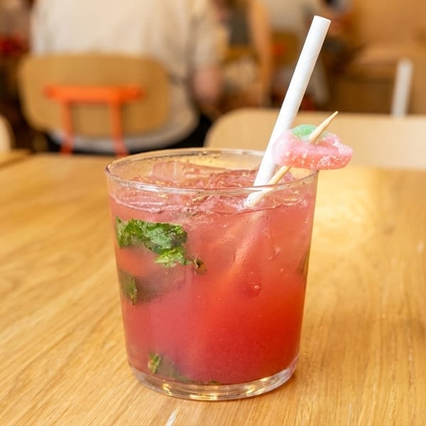 A vibrant pink cocktail with mint leaves, ice, a white straw, and a candy garnish on a wooden table.
