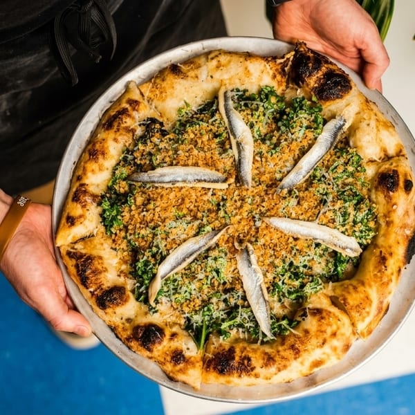 Hands holding a pizza topped with anchovies, breadcrumbs, and greens on a metal tray.