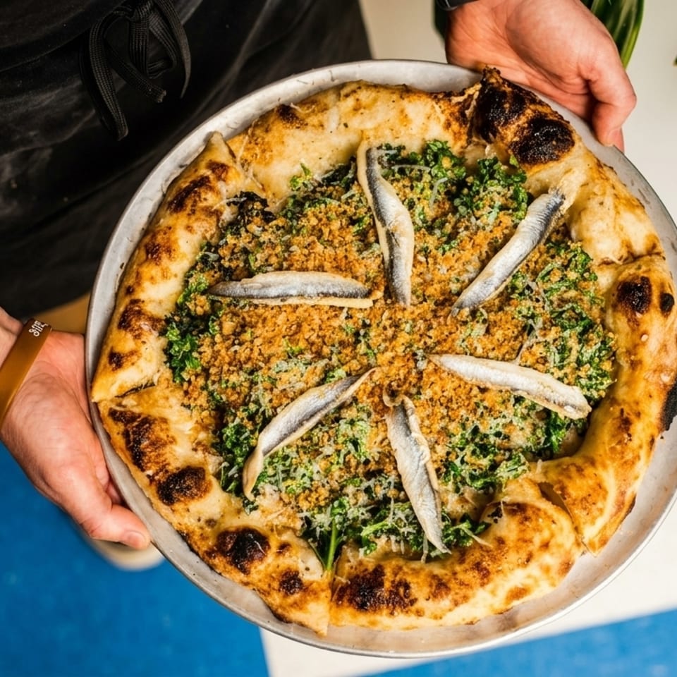 Hands holding a pizza topped with anchovies, breadcrumbs, and greens on a metal tray.